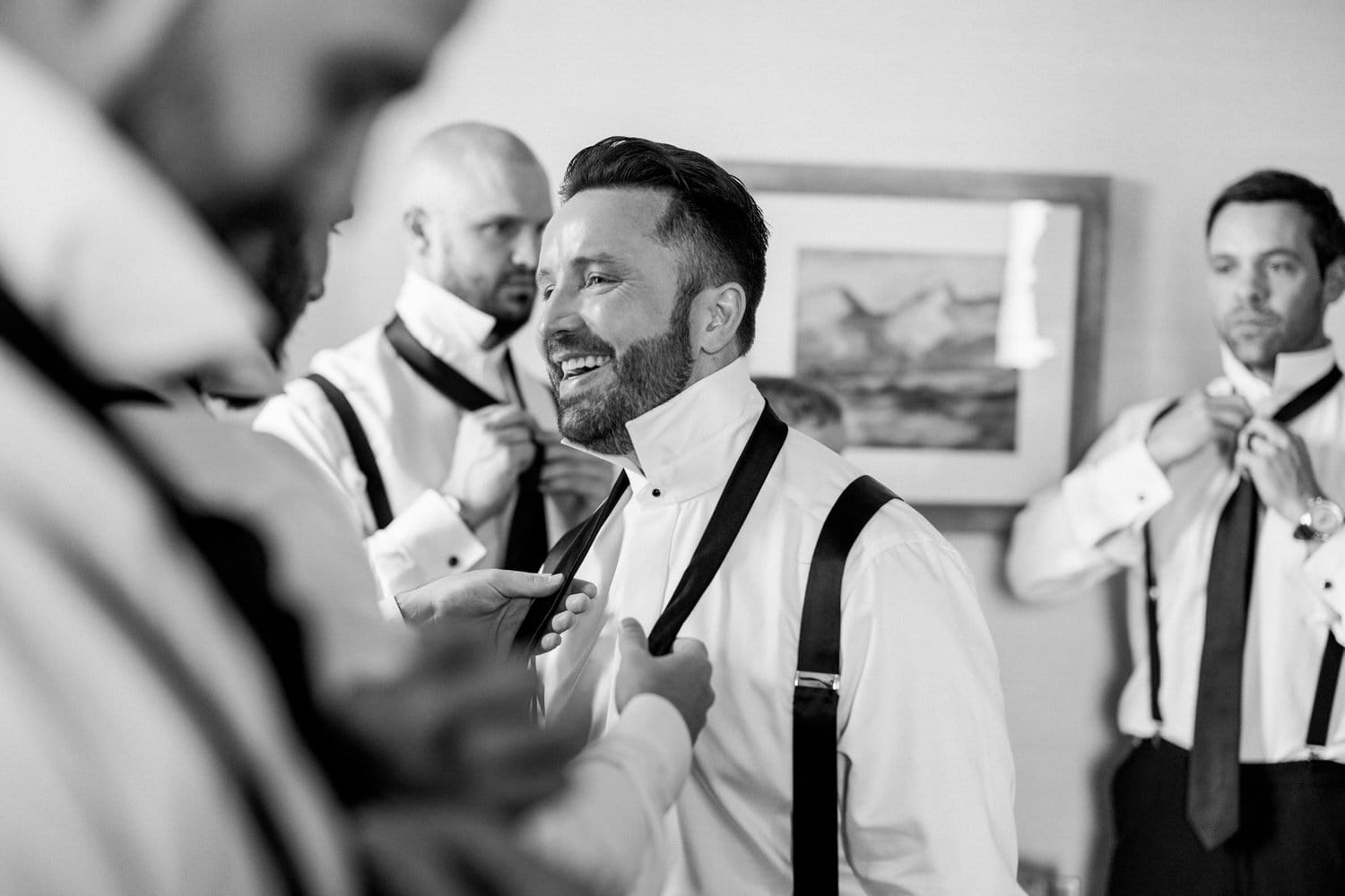 A cheerful groom smiles as he adjusts his outfit while surrounded by groomsmen preparing for a wedding, captured in a stylish black and white photo.