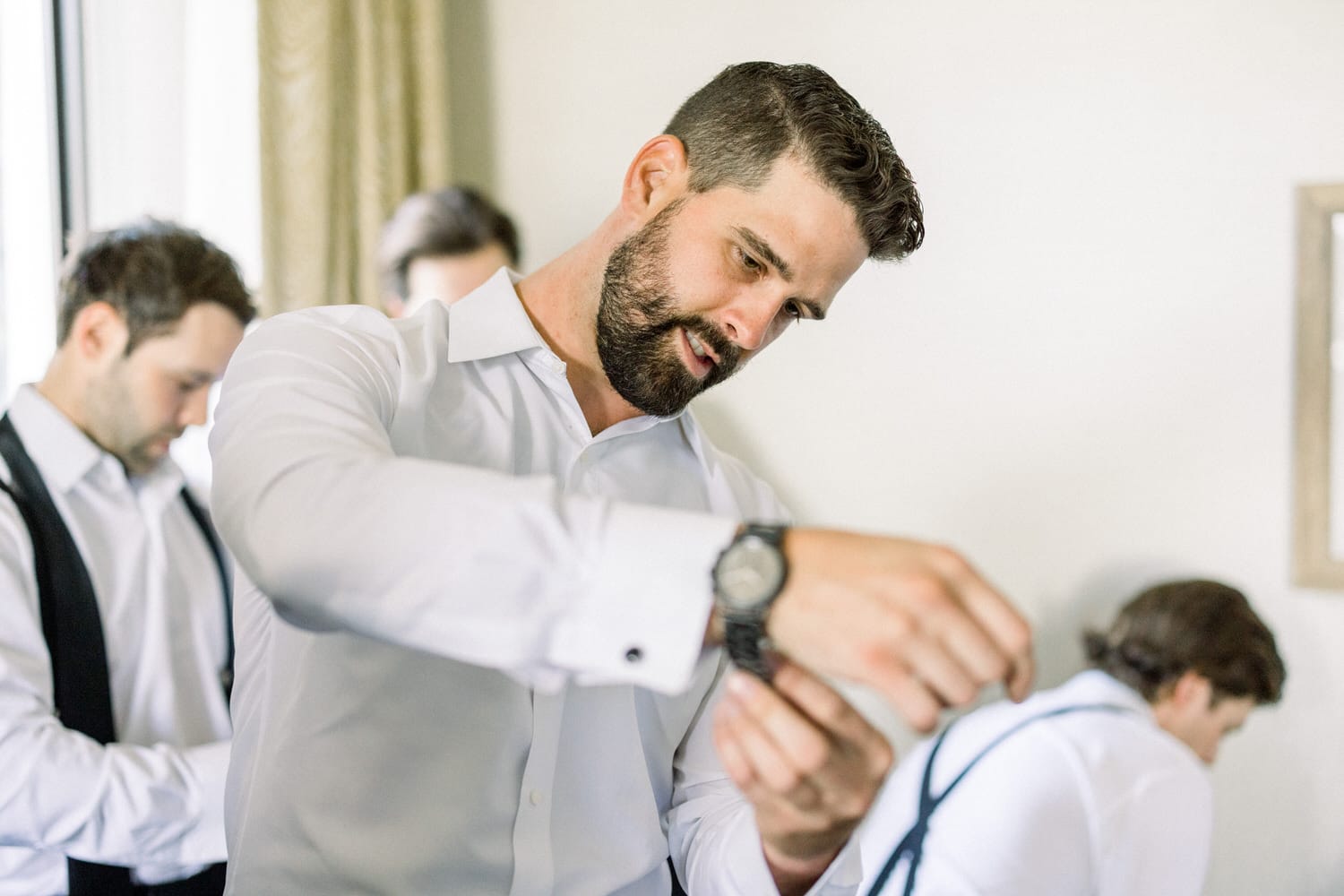 A man in a white dress shirt adjusts his cufflinks while two other men in the background prepare for a wedding.