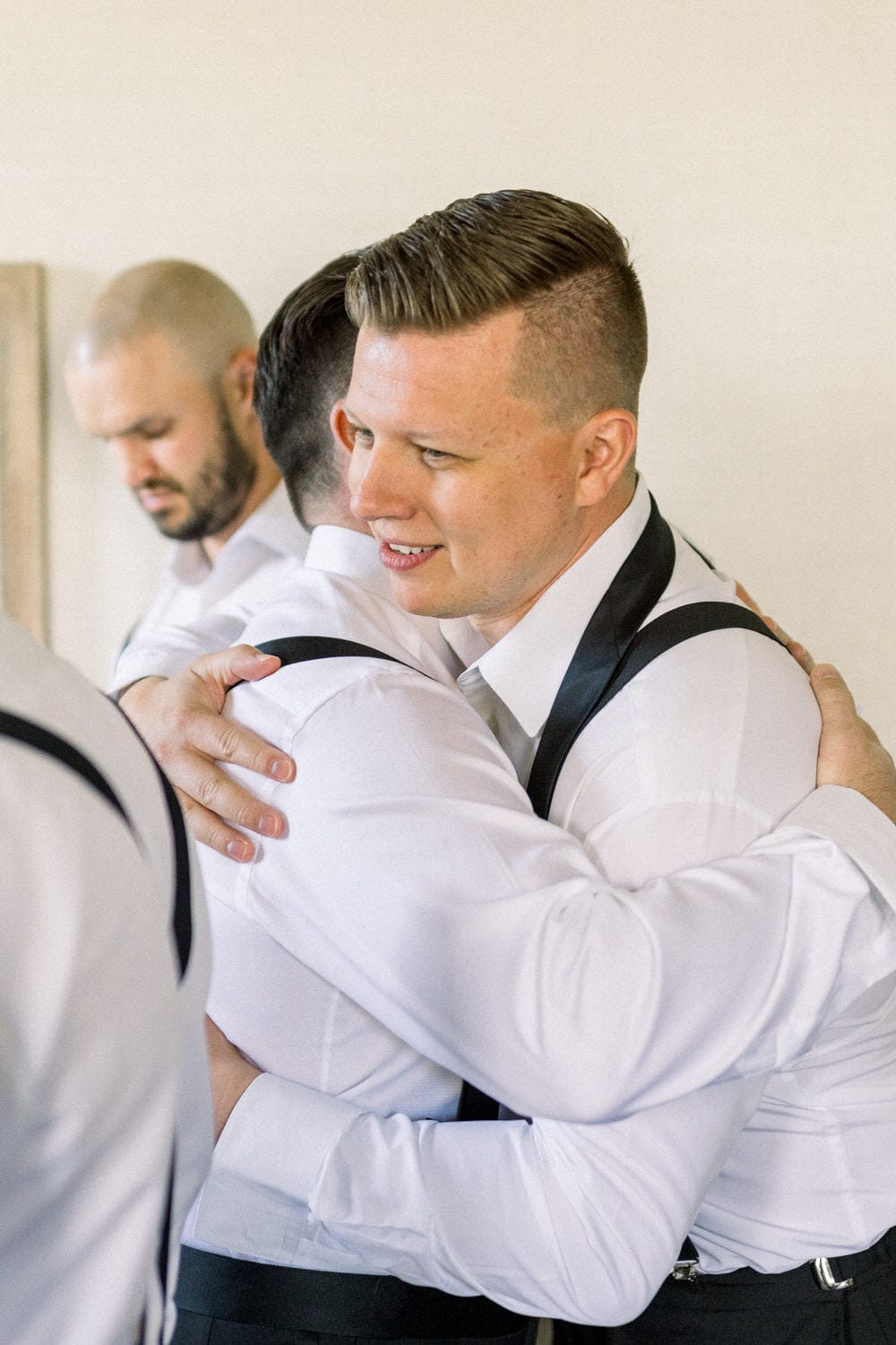 Two men in formal attire sharing an embrace, showcasing camaraderie and connection, with a third figure in the background.