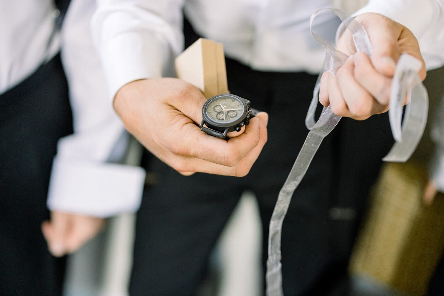 A close-up of a person holding a stylish black watch and a small gift box, with a ribbon in the other hand, dressed in formal attire.