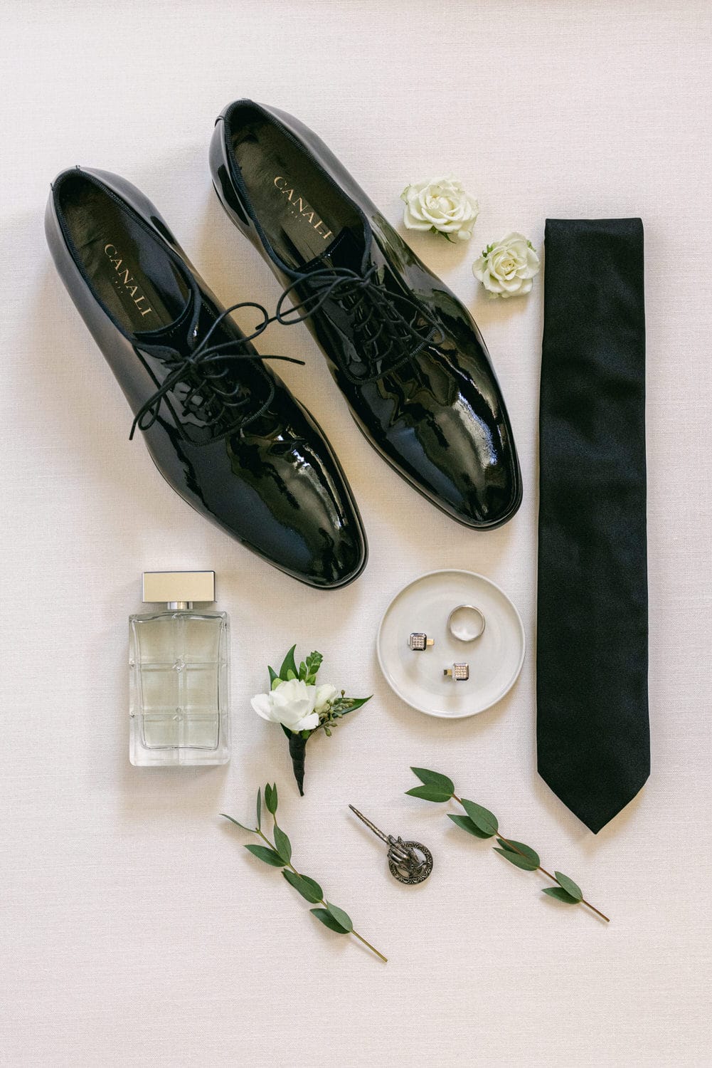 A stylish arrangement of black dress shoes, a tie, perfume, jewelry, and floral accents on a neutral background, perfect for a sophisticated outfit.