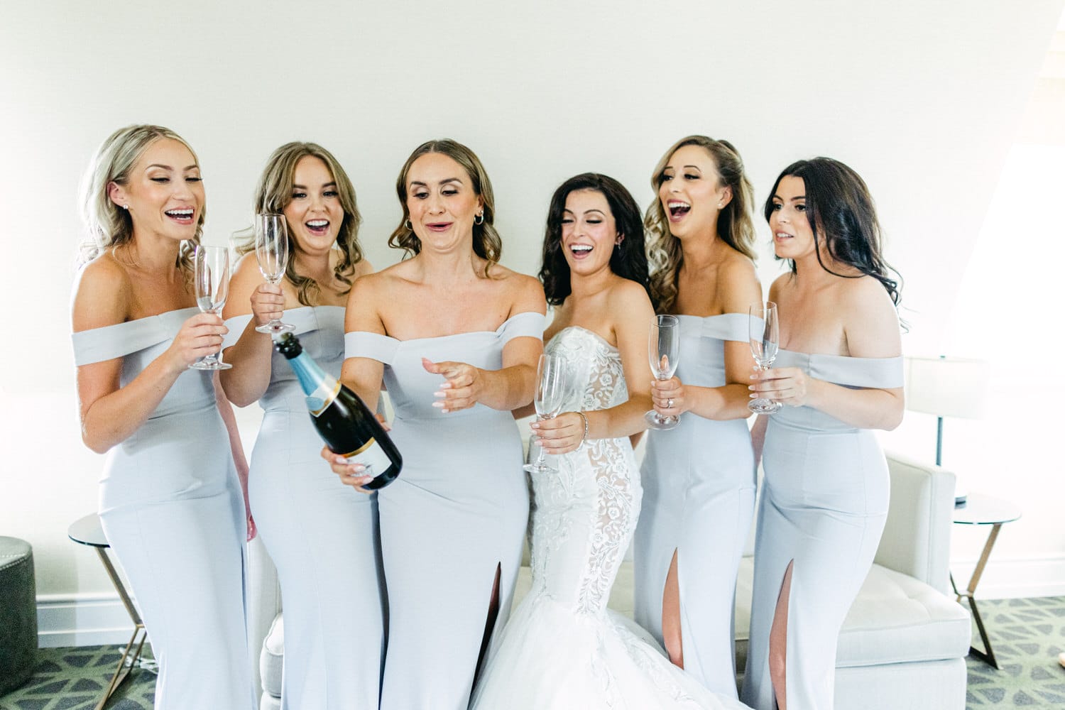 A joyful bridal party in grey dresses toasting and celebrating with champagne in a bright interior setting.