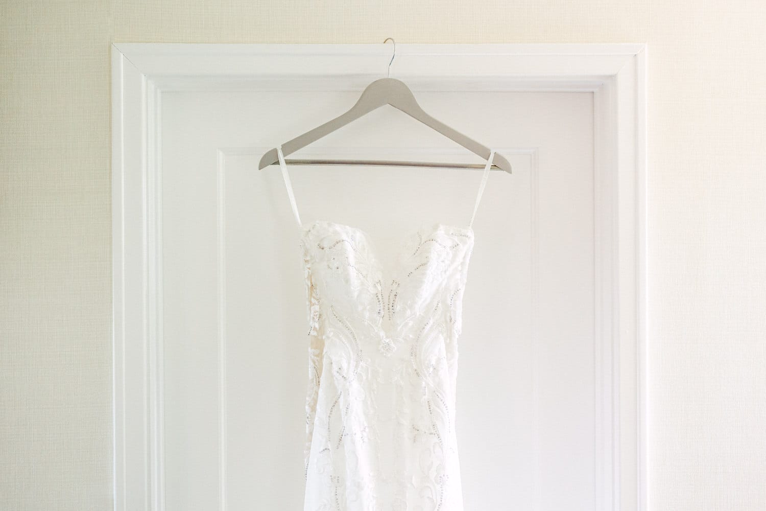 A beautifully crafted white wedding dress hanging on a hanger against a light-colored wall, showcasing intricate lace details and a delicate design.