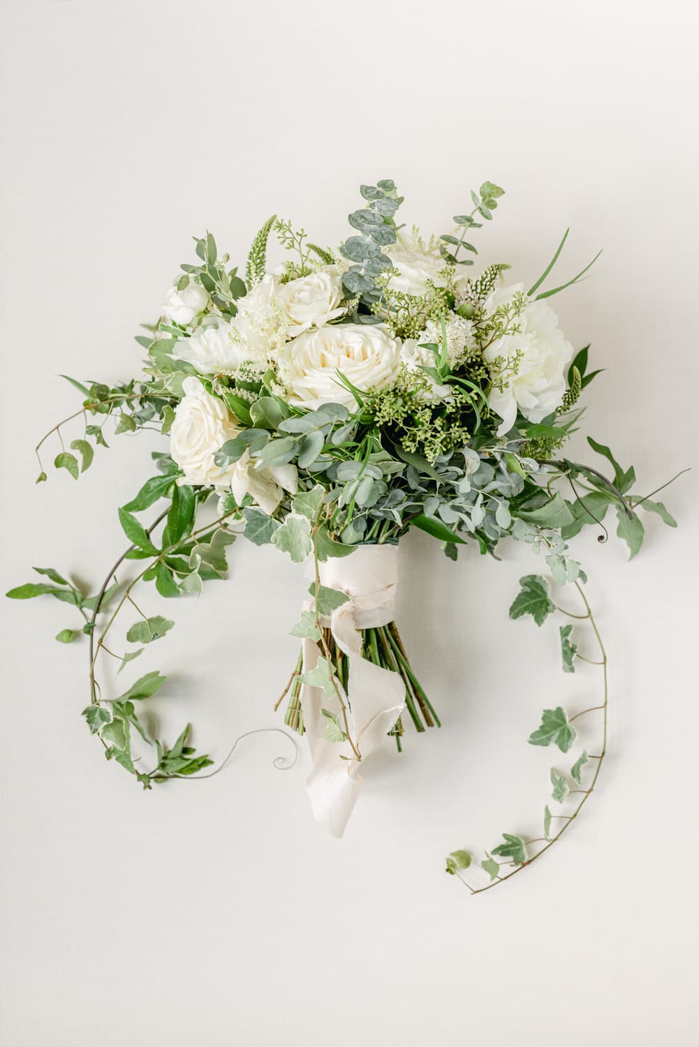 A beautiful arrangement featuring white roses, greenery, and delicate accents, artfully arranged and tied with ribbon against a soft, neutral background.