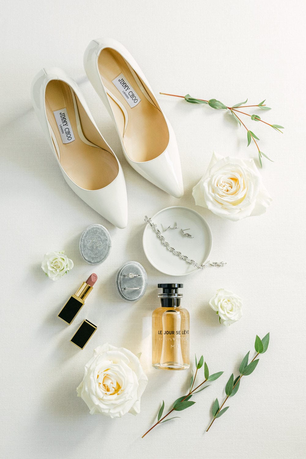 A flat lay of white high heels, lipstick, perfume, jewelry, and fresh flowers arranged beautifully on a textured surface.