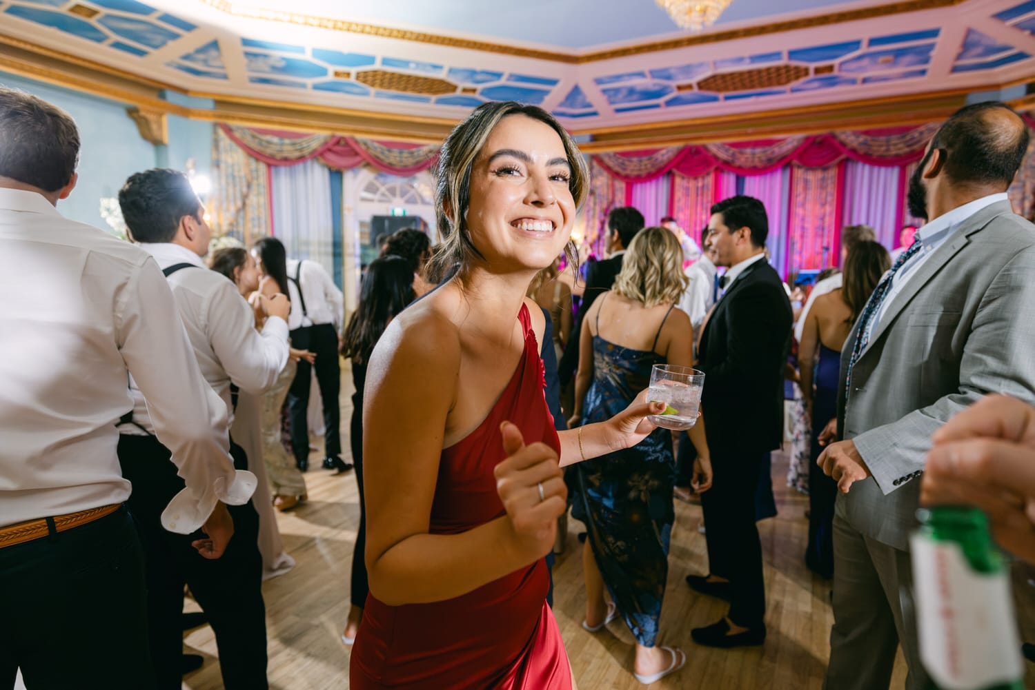 A smiling woman in a red dress enjoying a vibrant party, holding a drink with guests dancing in the background.