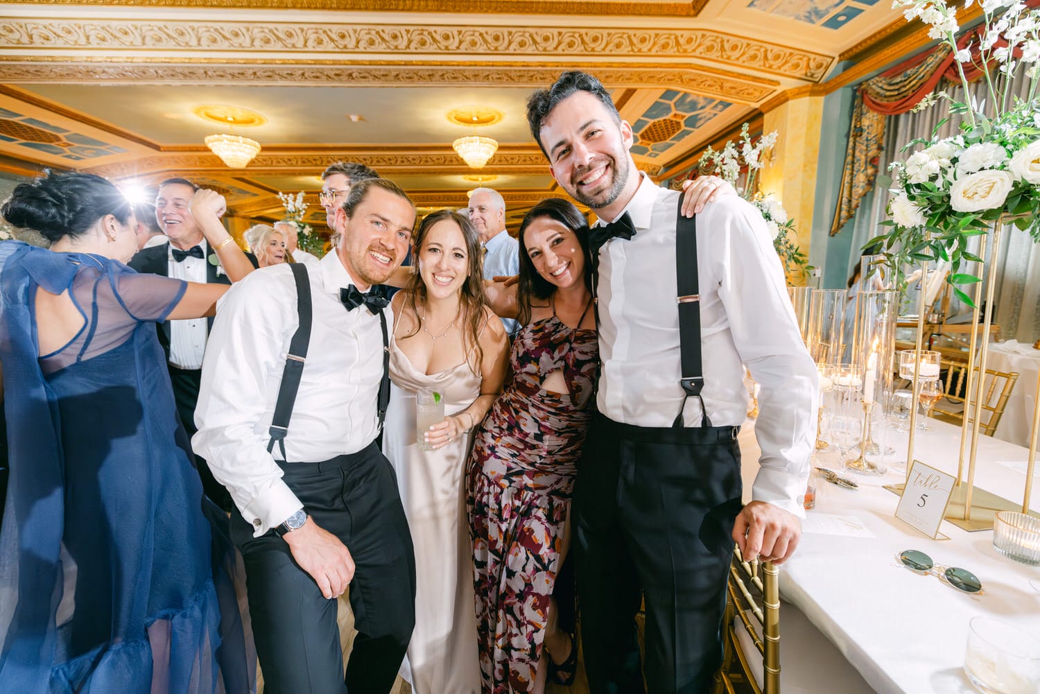 Group of friends smiling and posing together at a lively wedding celebration with elegant decor in the background.