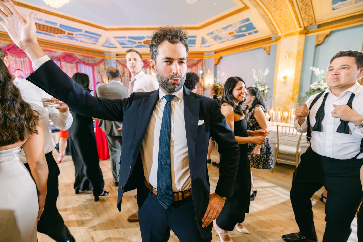 A well-dressed man poses dramatically while dancing at a lively celebration, surrounded by guests in elegant attire.