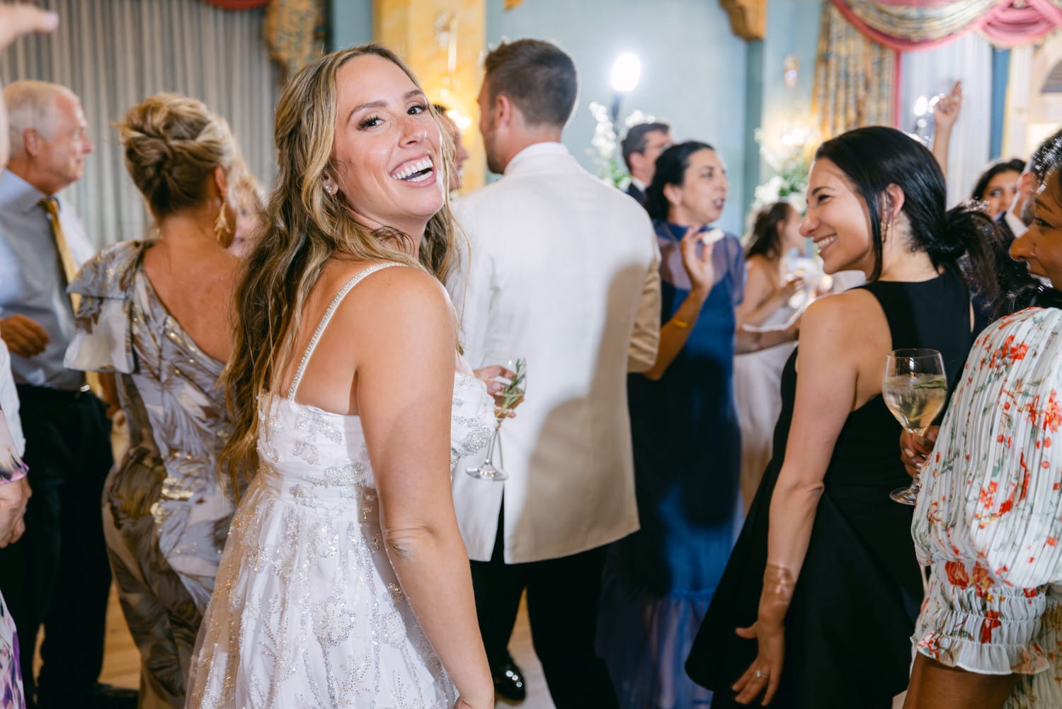 A smiling woman in a sparkling white dress enjoys the festivities at a lively wedding reception, with guests dancing and socializing in the background.