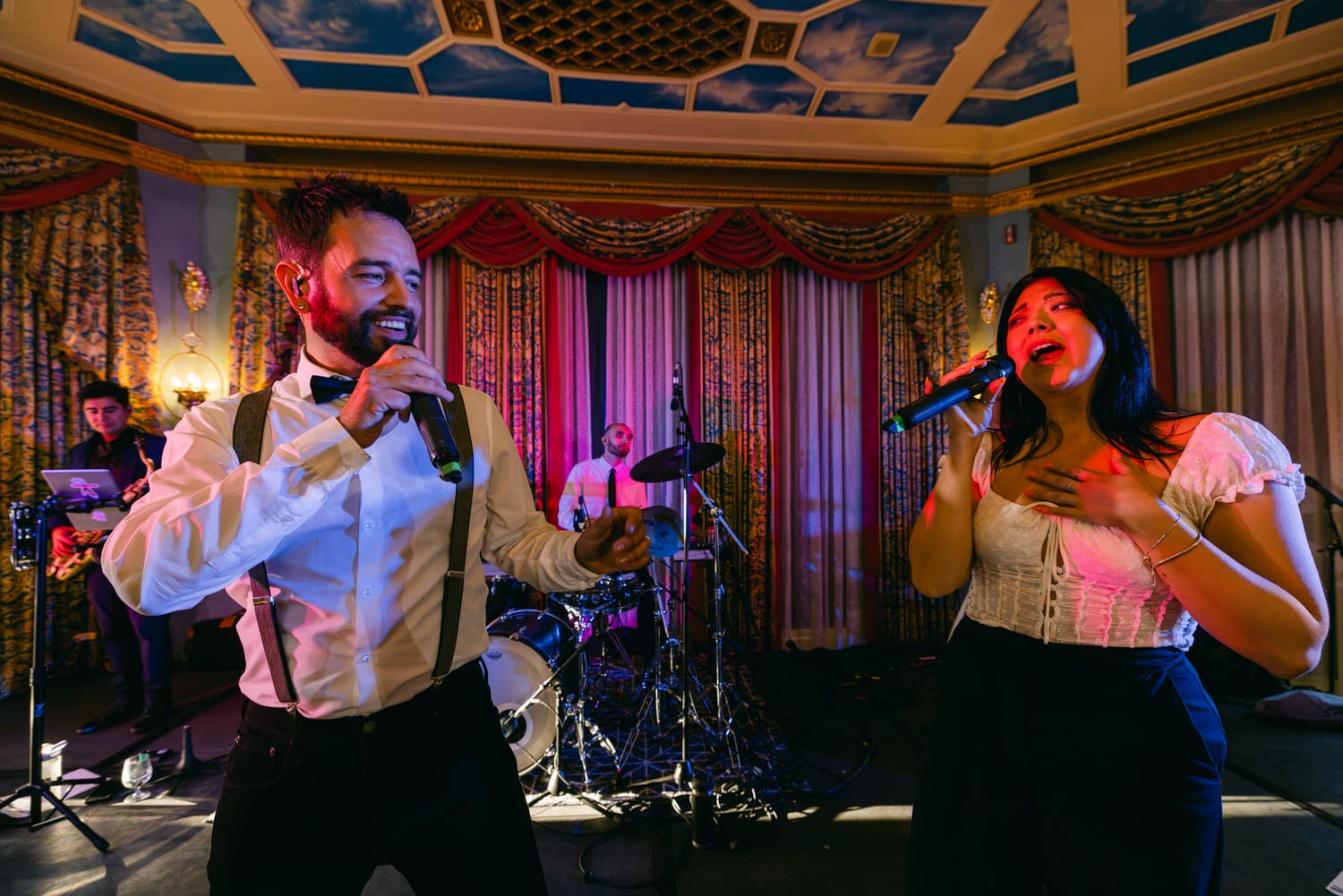 Live Music Performance::A male and female singer performing on stage with a live band in a well-decorated venue.