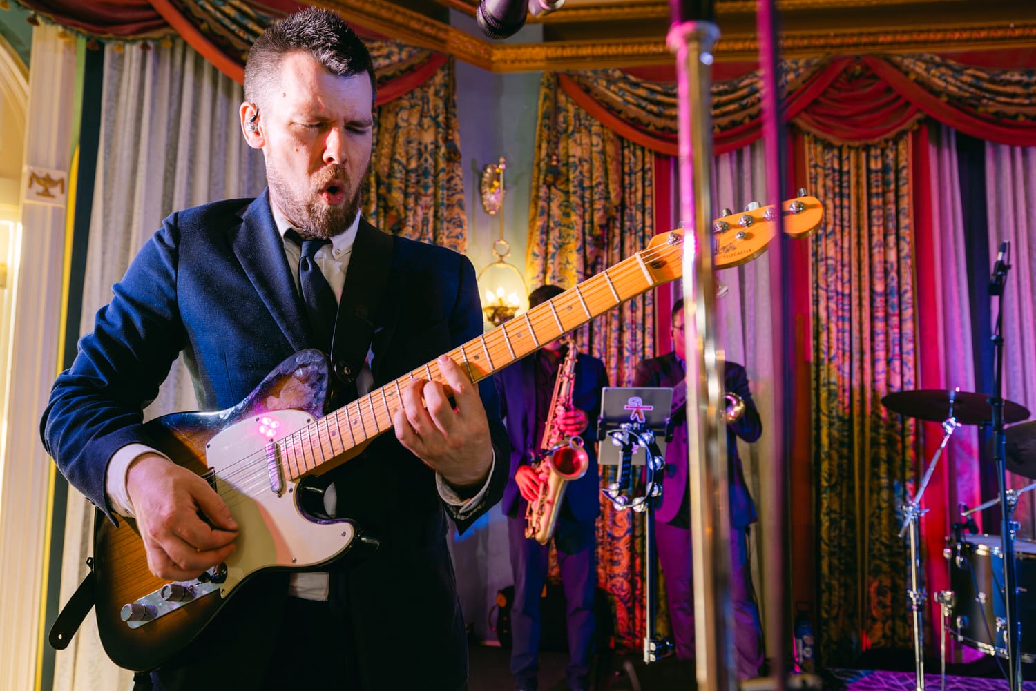 Live Music Performance:::A musician passionately plays the electric guitar on stage, surrounded by colorful curtains and band members in the background.