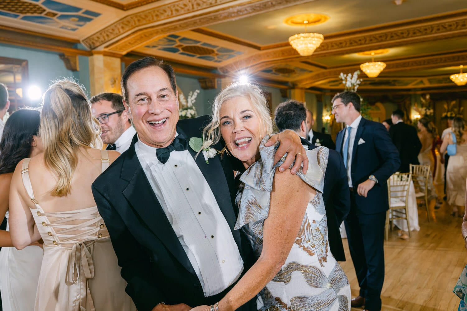 A happy couple poses for a photo while celebrating at an elegant event, surrounded by guests and a beautifully decorated venue.