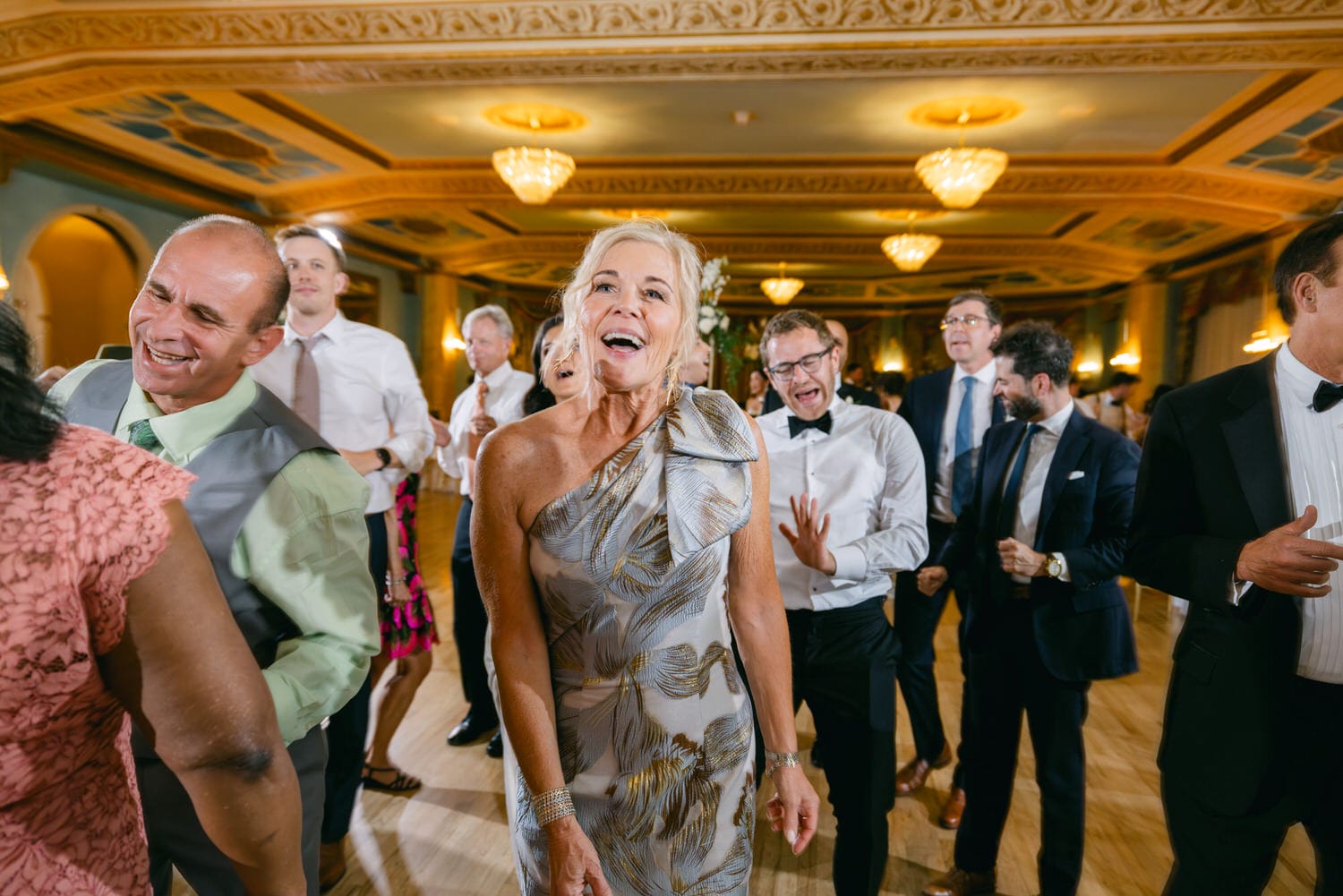 A joyful group of guests dancing together at a formal event, with one woman in a stylish silver dress smiling brightly in the foreground.