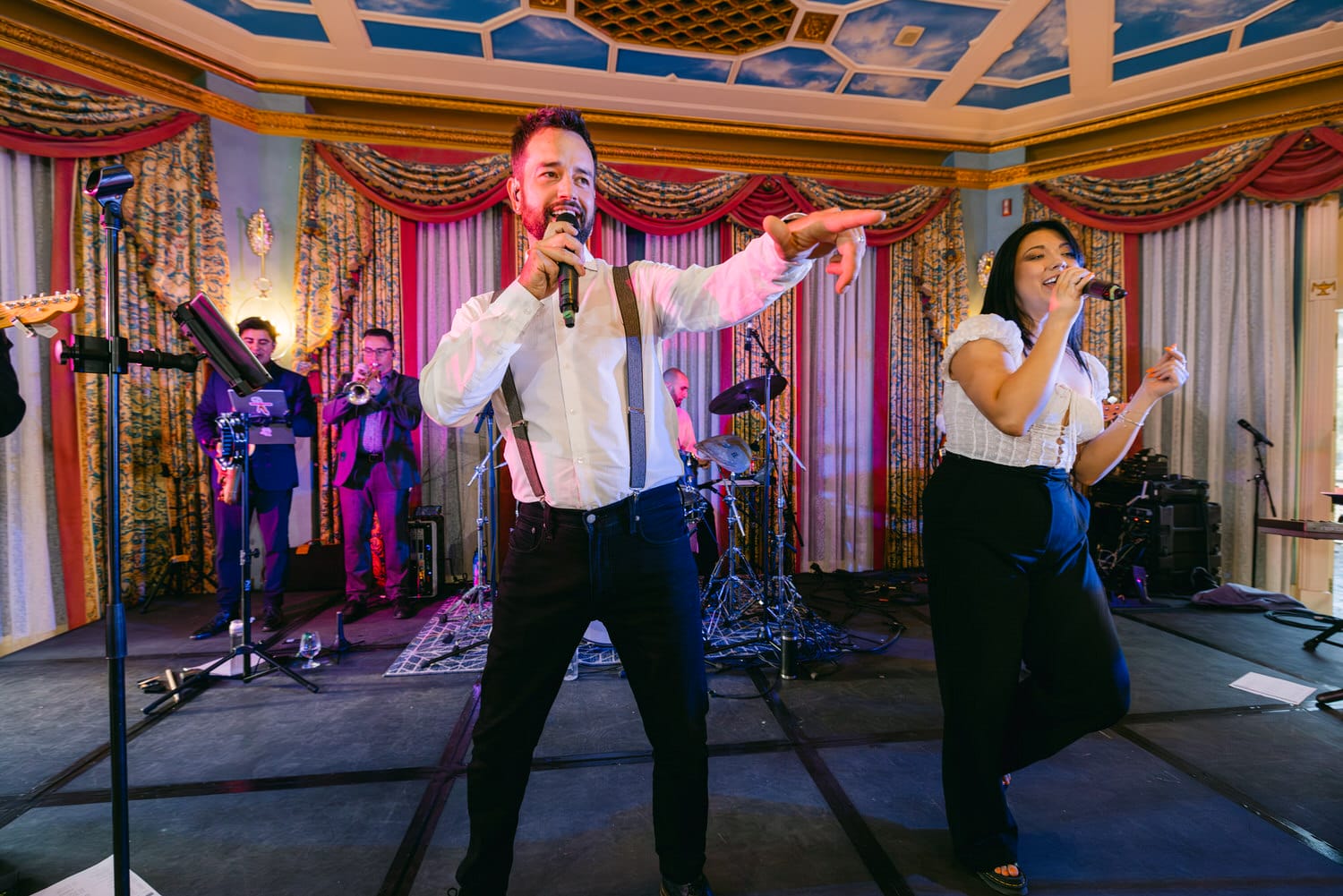 A lively music performance featuring a male vocalist engaging the audience, accompanied by a female singer, with a band in the background.