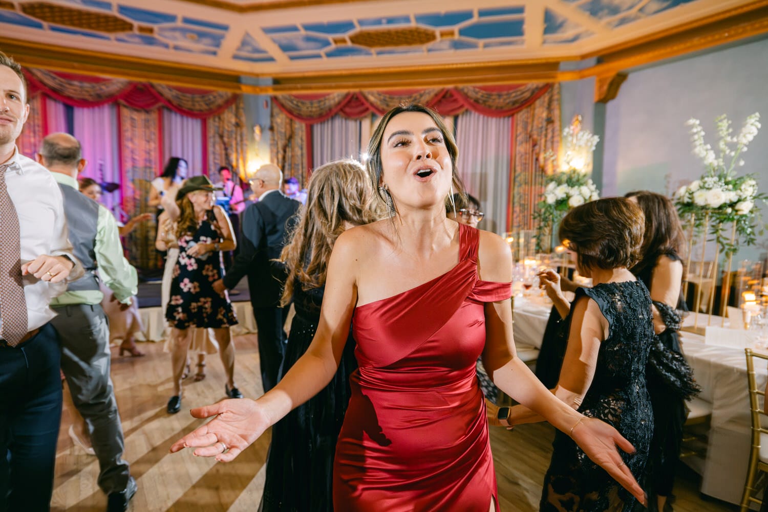 A woman in a red dress joyfully dances amidst a lively wedding reception, surrounded by guests at tables and enjoying the music.