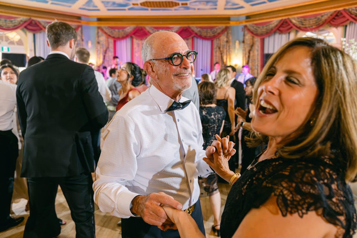 A smiling older couple dances hand in hand at a lively event, surrounded by guests enjoying the festivities in an elegantly decorated venue.