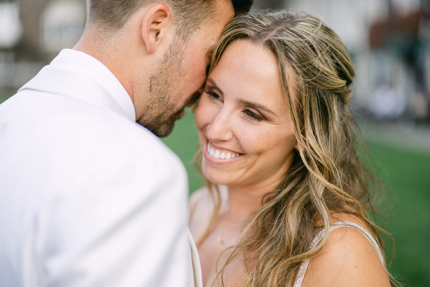 A couple sharing a tender moment, with the man whispering in the woman's ear, both smiling warmly against a soft-focus outdoor background.