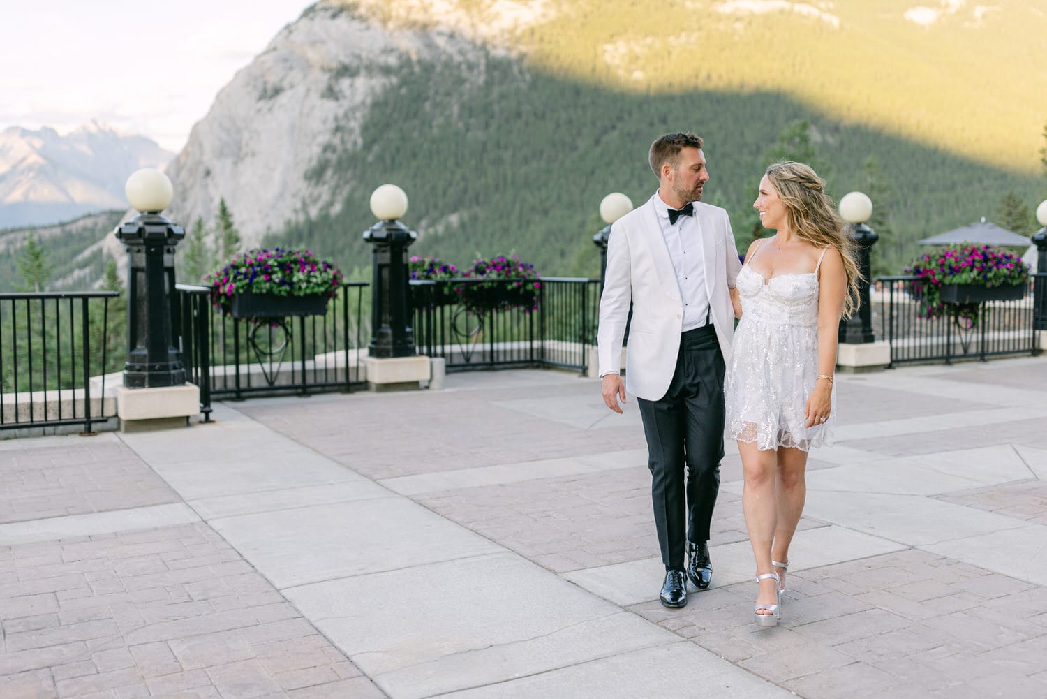 A couple dressed elegantly walks hand in hand on a scenic outdoor terrace with mountains in the background and floral arrangements.