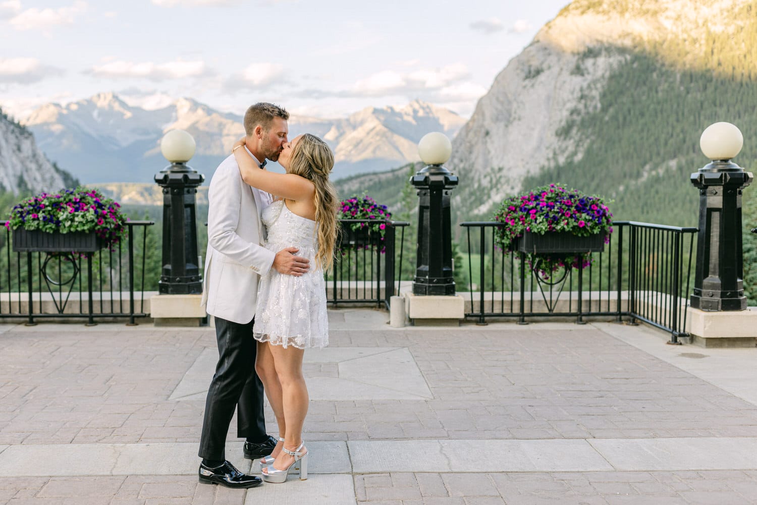 A couple sharing a kiss against a breathtaking mountain backdrop, adorned with purple flowers and a clear sky.