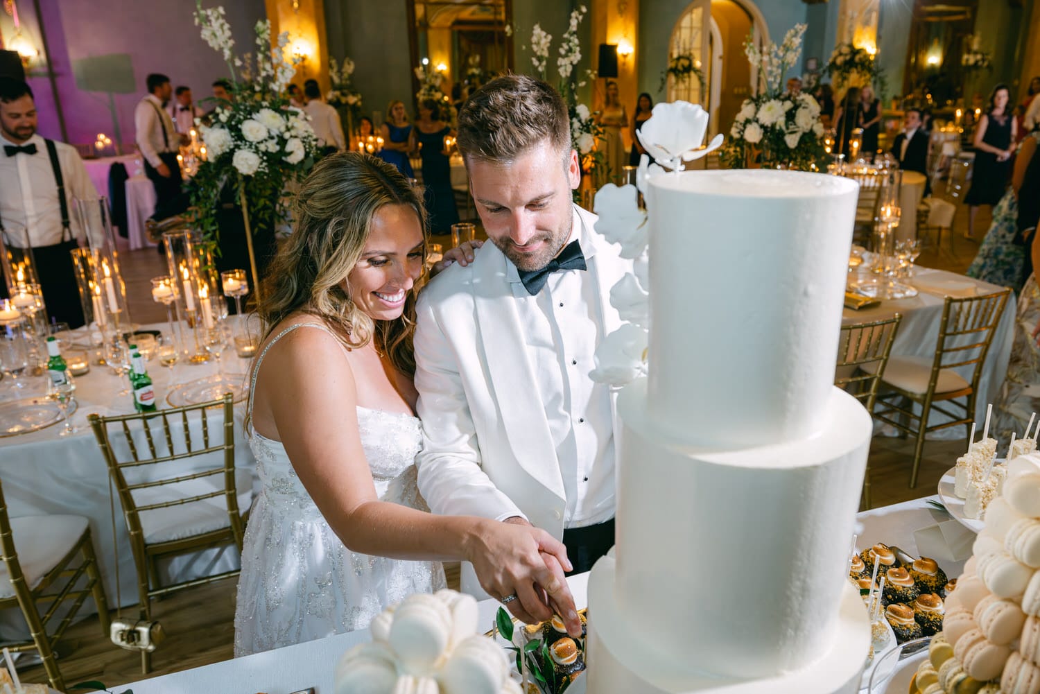 A joyful couple in formal attire slicing a wedding cake surrounded by elegant decor and guests celebrating in the background.