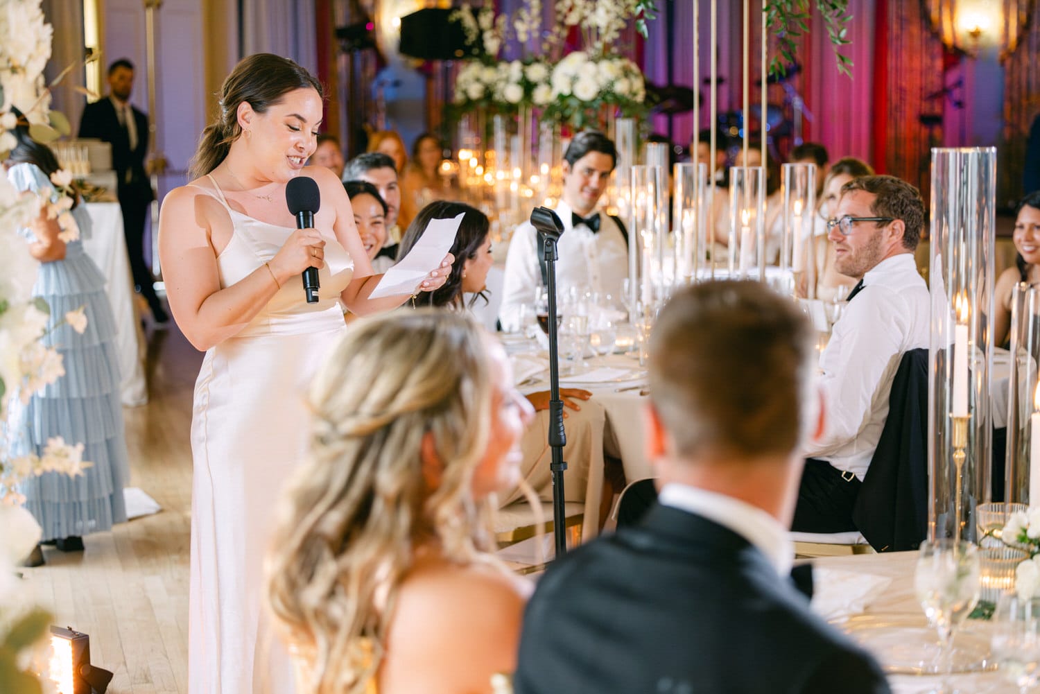 A woman delivers a heartfelt toast at a wedding reception, with guests listening attentively in an elegantly decorated venue.