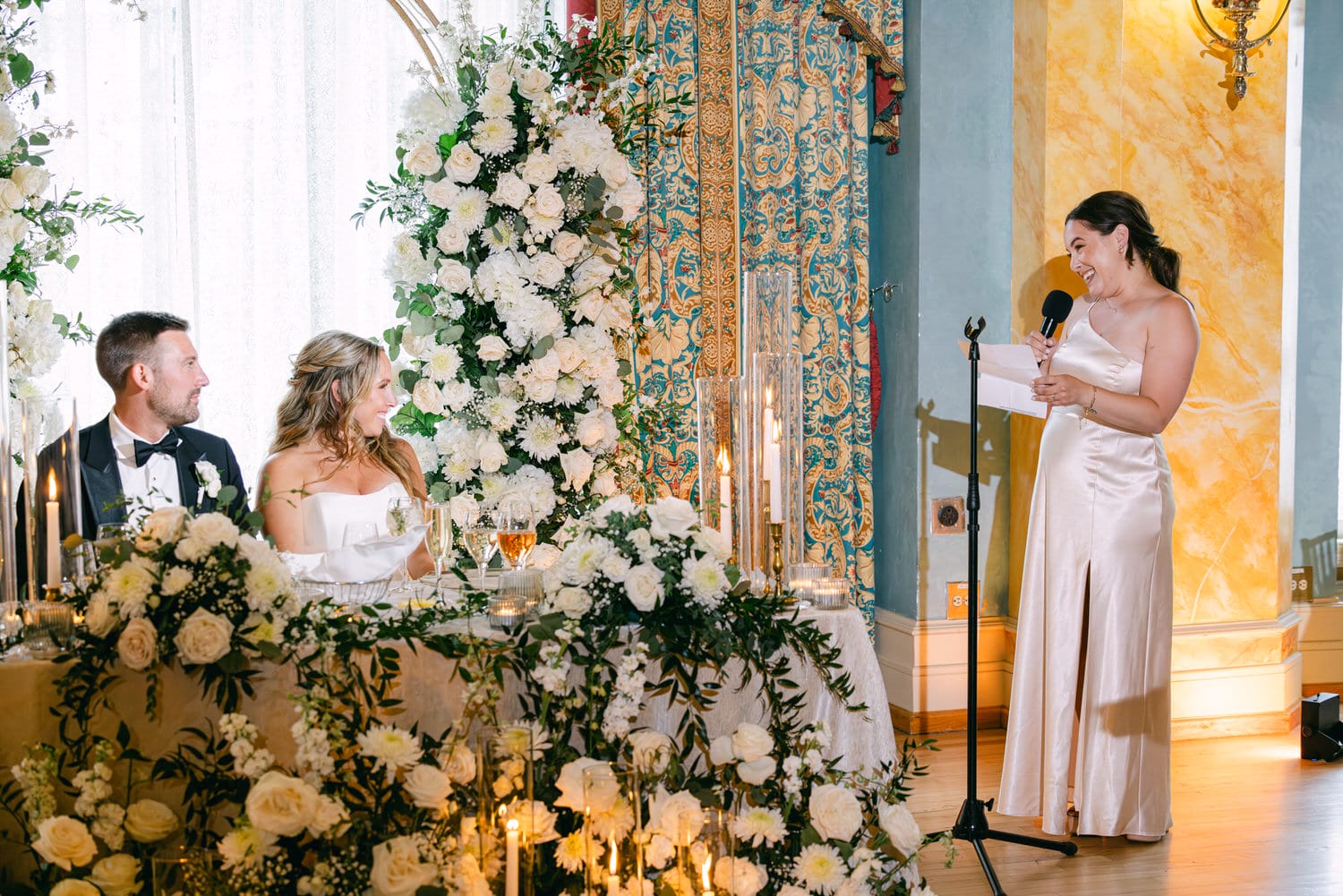 A woman in a satin gown delivers a toast at a wedding reception, while the couple sits at a beautifully decorated table surrounded by floral arrangements and candles.