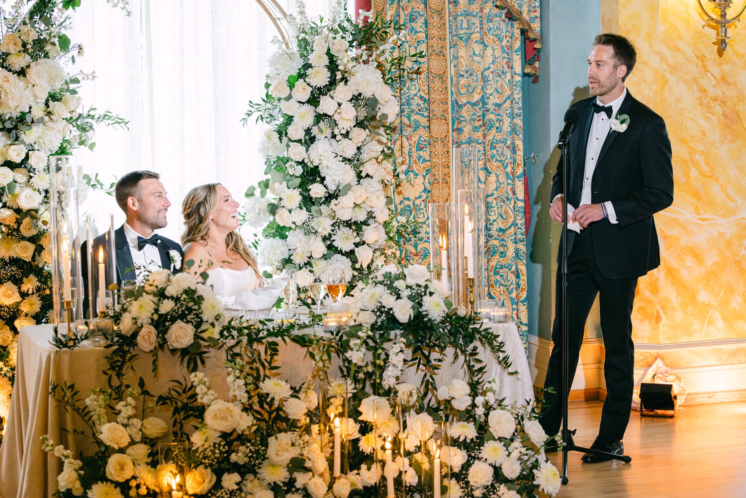 Happy couple listening to a heartfelt toast amidst elegant floral decorations at their wedding reception.