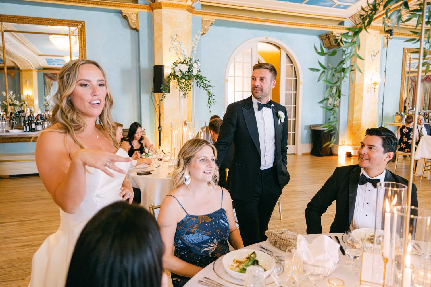 A bride shares a heartfelt moment while speaking to guests at a beautifully decorated reception, with elegant dining settings and a warm atmosphere.