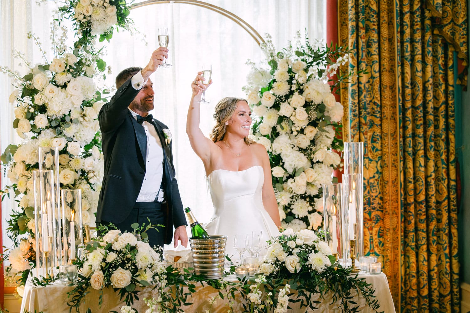 A bride and groom toast with champagne glasses at their wedding reception, surrounded by elegant floral arrangements.