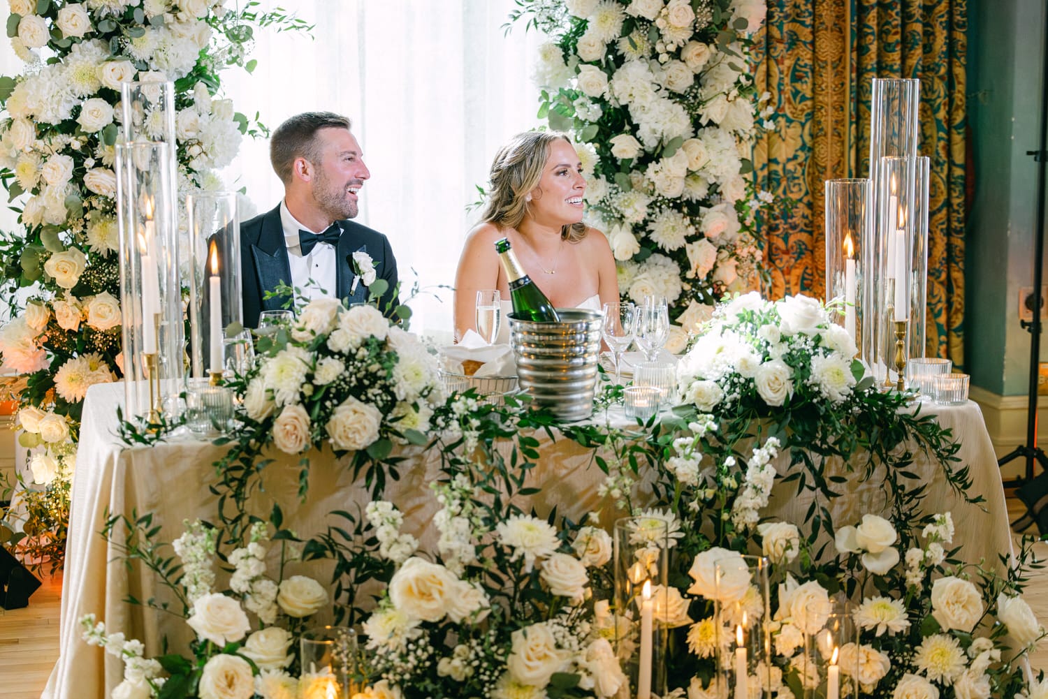 A smiling bride and groom seated at a beautifully decorated head table adorned with white flowers and candles during their wedding reception.