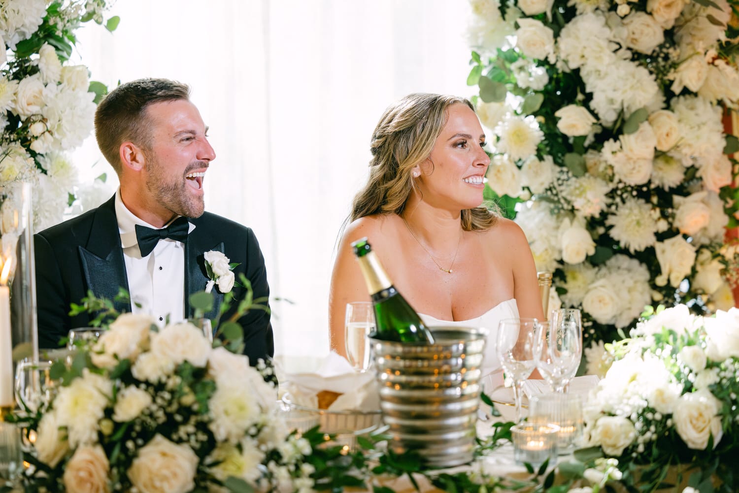 A newlywed couple smiling and enjoying their reception, surrounded by elegant floral arrangements and champagne on the table.