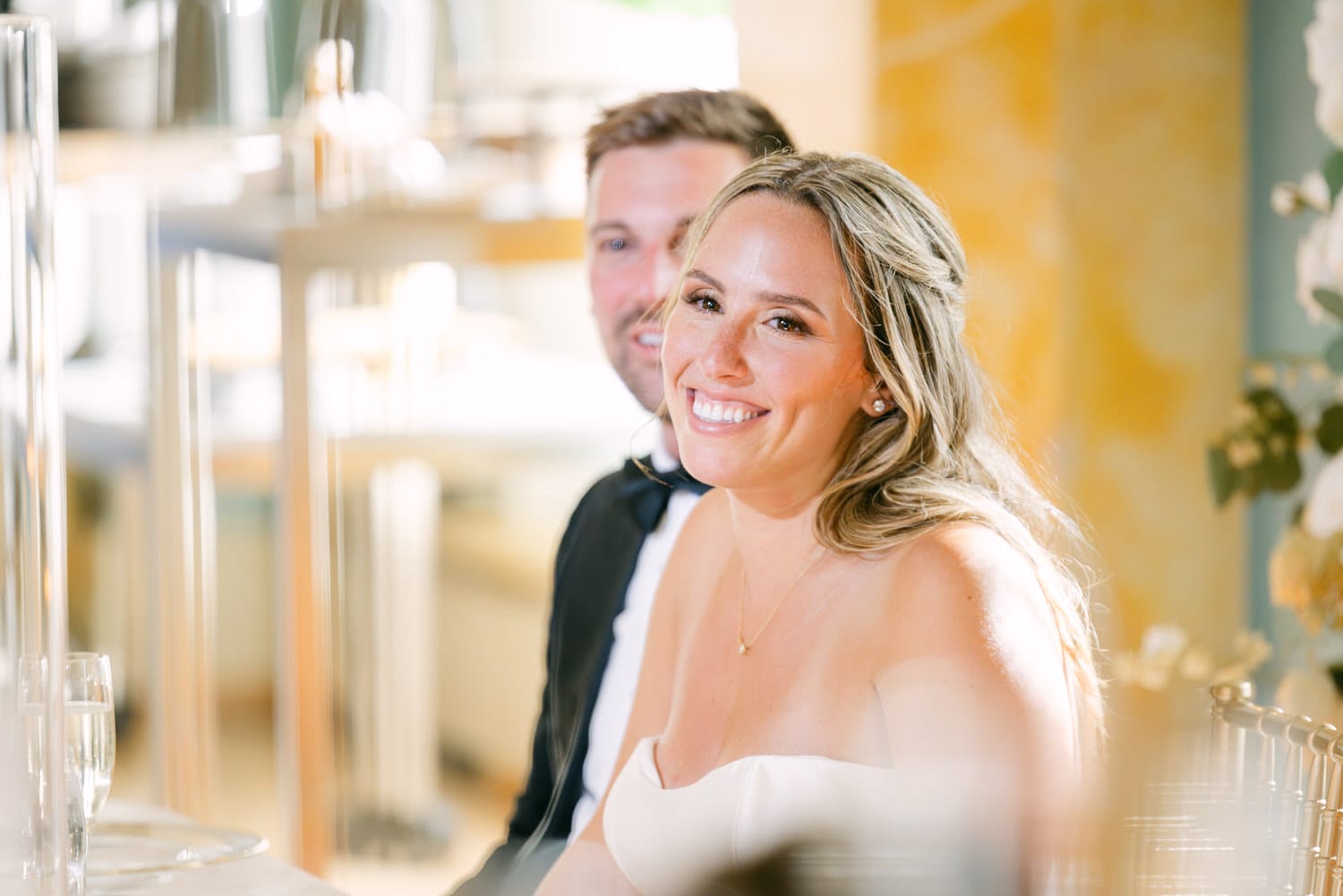 A smiling woman in a strapless dress sits at a table during a celebration, with a blurred figure in the background.