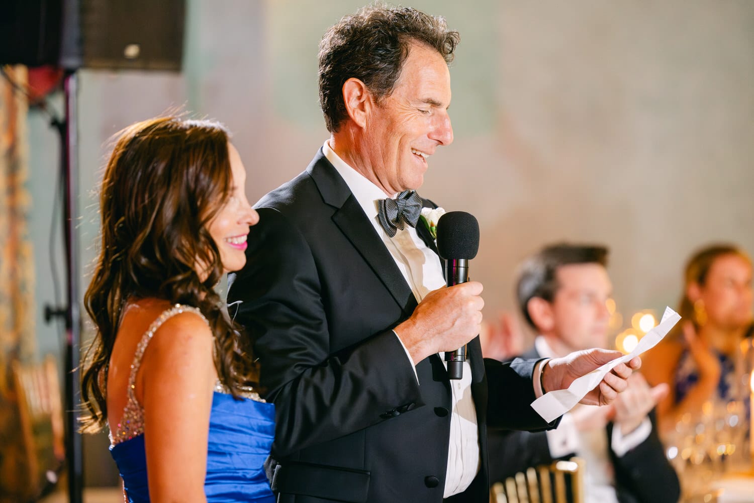 A man in a tuxedo delivers a heartfelt speech while a woman in a blue dress smiles beside him, capturing a memorable moment at a lively event.