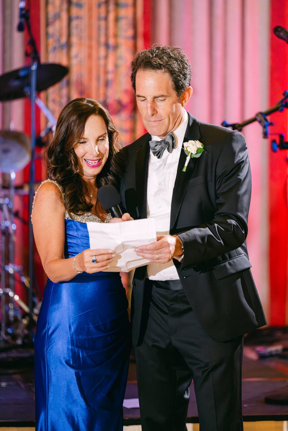 A couple at a festive event, smiling while reading from a paper, with musical instruments in the background.