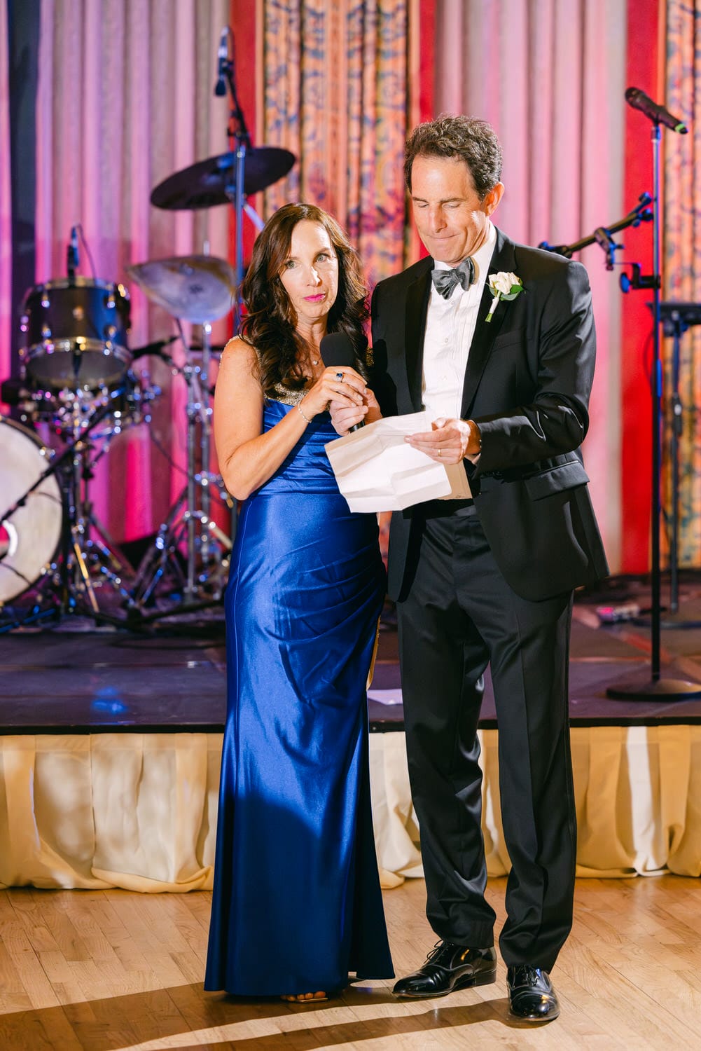 A man and woman, dressed in formal attire, stand together at a microphone while the woman holds a speech paper, with musical instruments in the background.