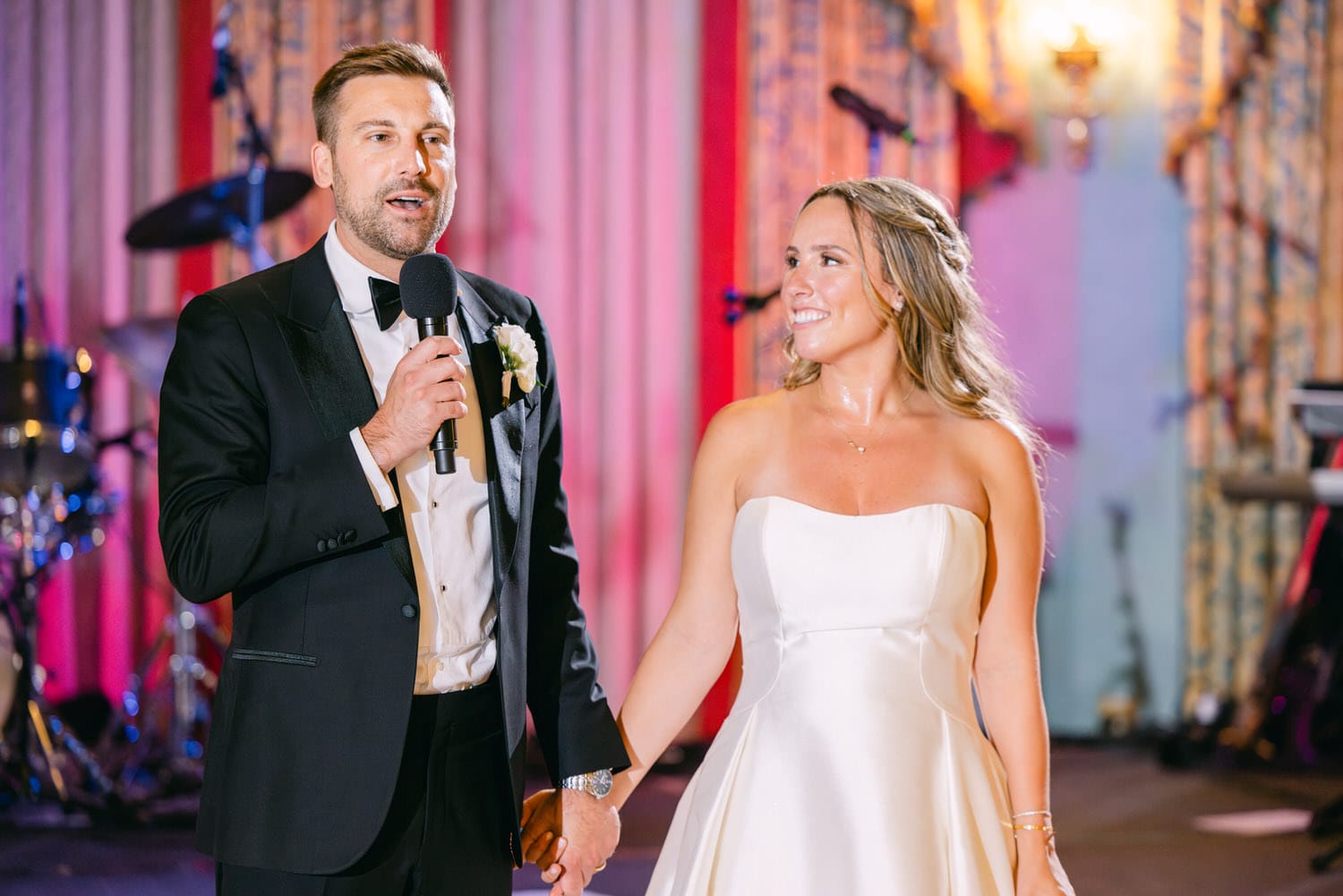 A groom delivers a heartfelt speech while holding hands with his bride, both smiling at each other during their wedding reception.