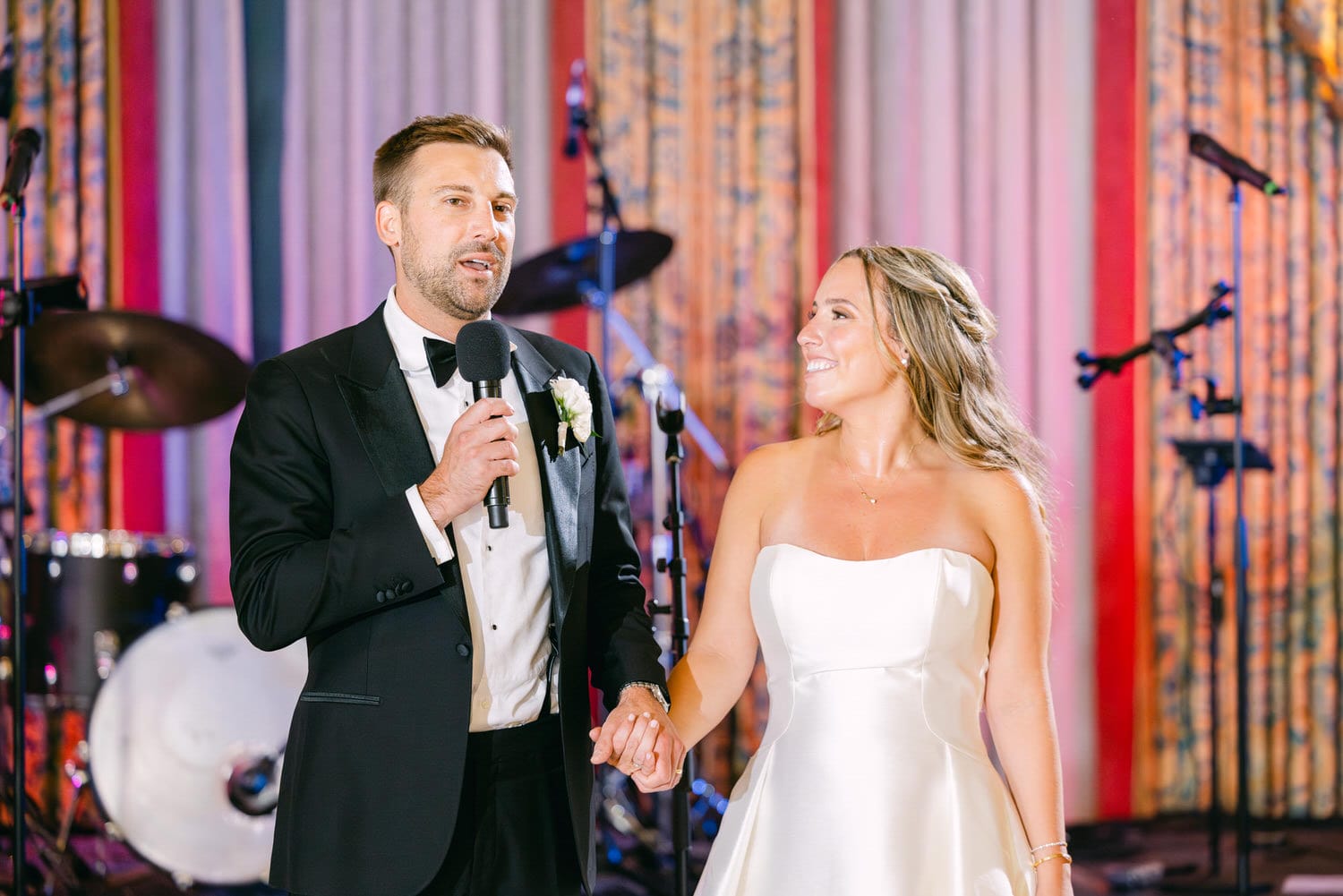 A man in a tuxedo delivers a heartfelt toast while holding hands with a smiling woman in a wedding dress, surrounded by a festive backdrop with musical instruments.