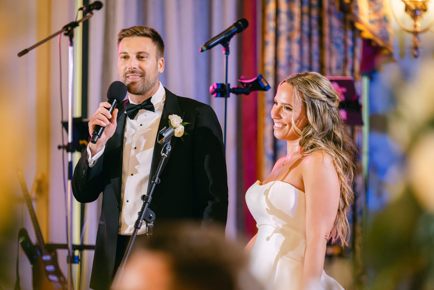 A groom giving a heartfelt speech while the bride smiles, with wedding decorations in the background.