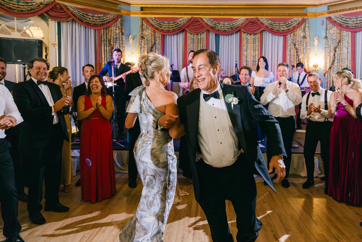 A joyful couple dances at a festive event, surrounded by happy guests clapping and enjoying the live band in an elegantly decorated hall.