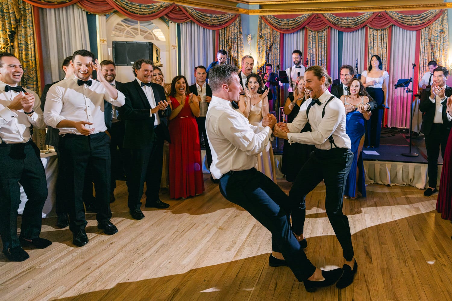 Two men engaging in a lively dance as a joyful crowd cheers around them during a festive event.