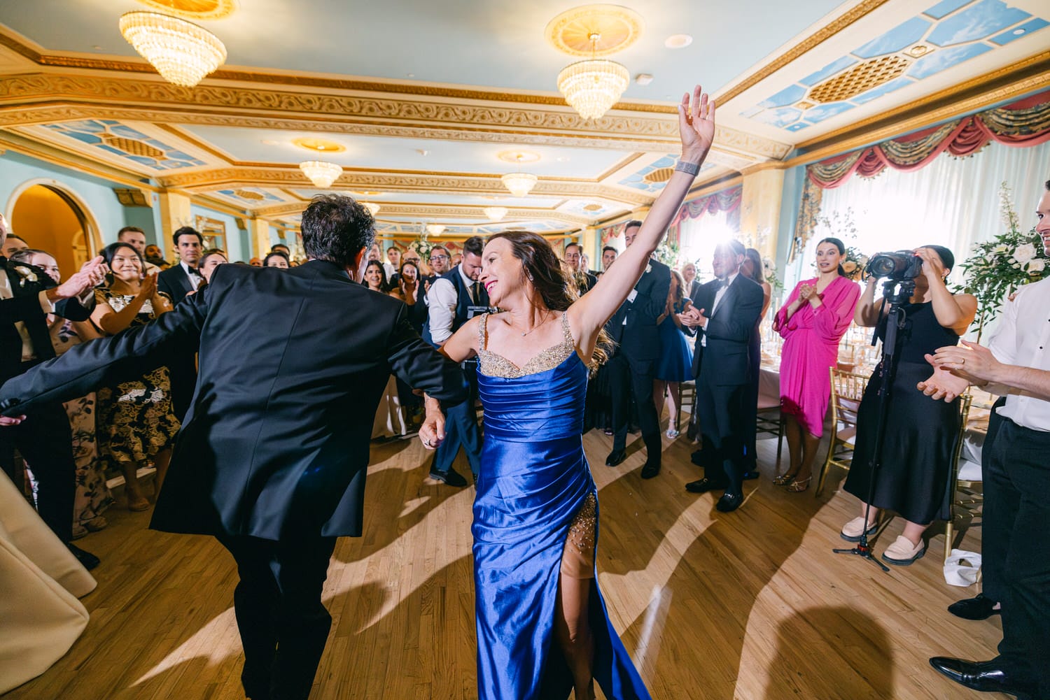 A joyful couple dances in a luxurious ballroom surrounded by cheering guests, capturing the festive atmosphere of a wedding celebration.