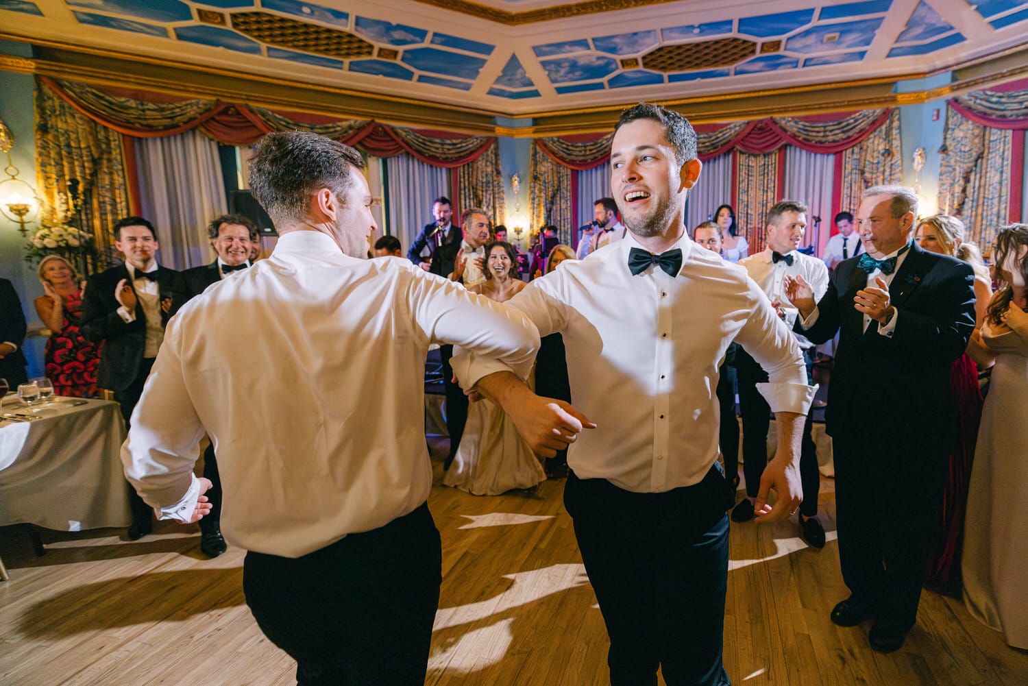 Two men dancing joyfully at a festive event, surrounded by an engaged audience in an elegantly decorated venue.