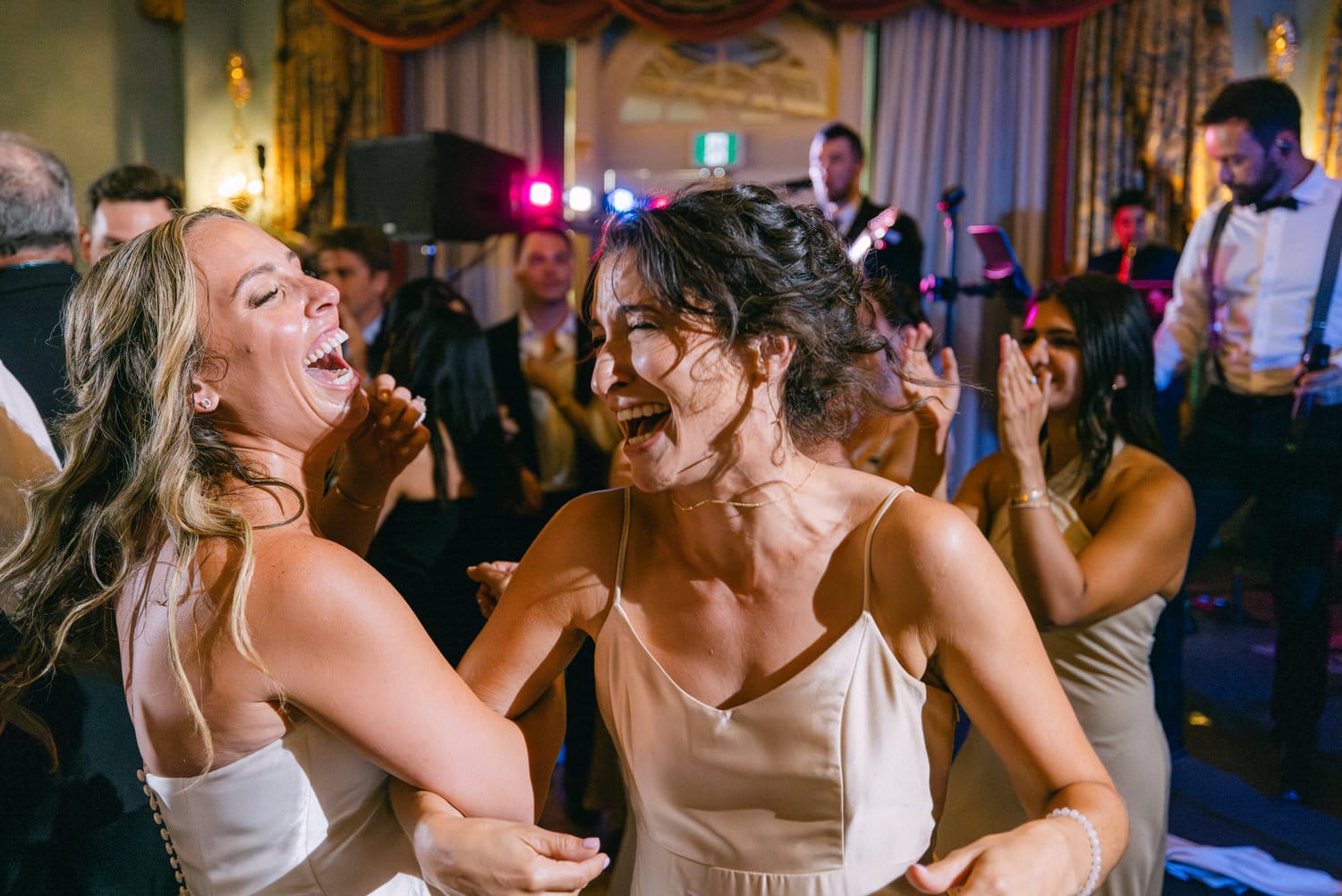 Two women laughing and dancing together at a lively event, surrounded by friends and colorful lights.