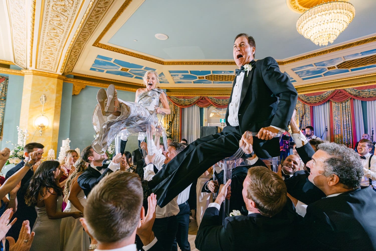 A lively wedding scene featuring a man and woman joyfully lifted on chairs by their guests, surrounded by a crowd celebrating in an elegant venue.