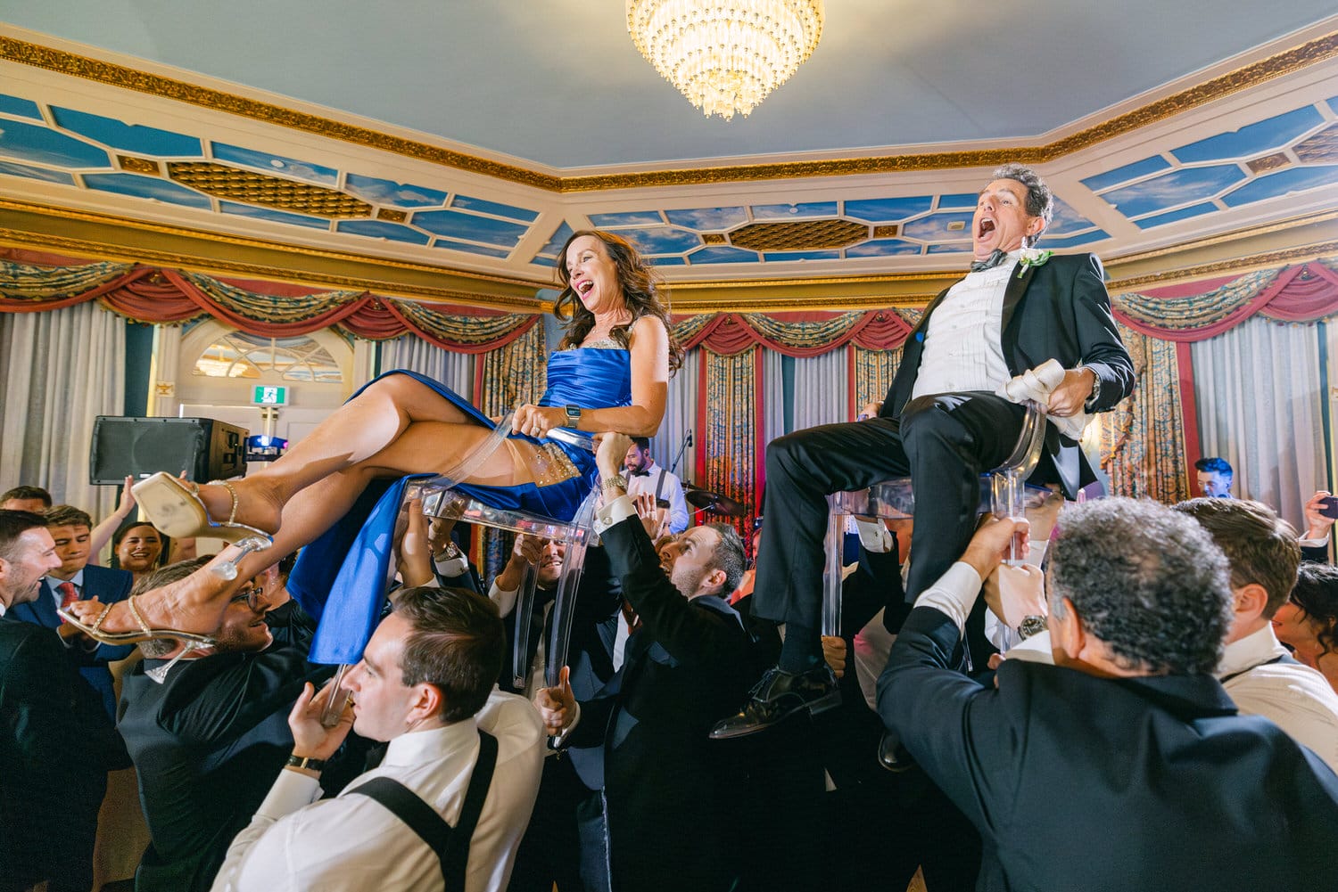 A joyful couple being lifted in transparent chairs by guests during a lively wedding celebration in an elegant venue.