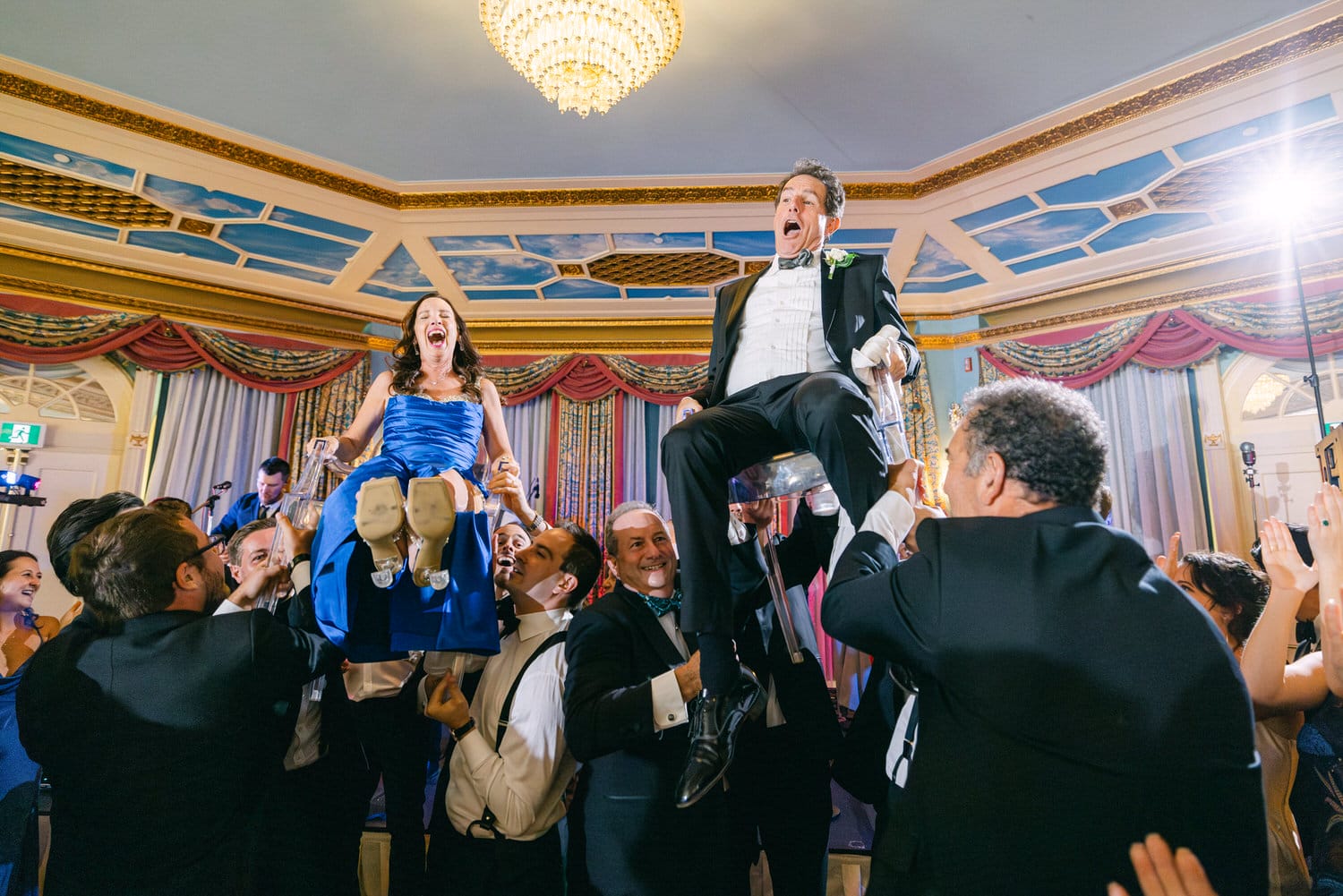 A couple is joyfully lifted by their friends during a wedding reception, surrounded by laughter and celebration, set in an elegant venue.