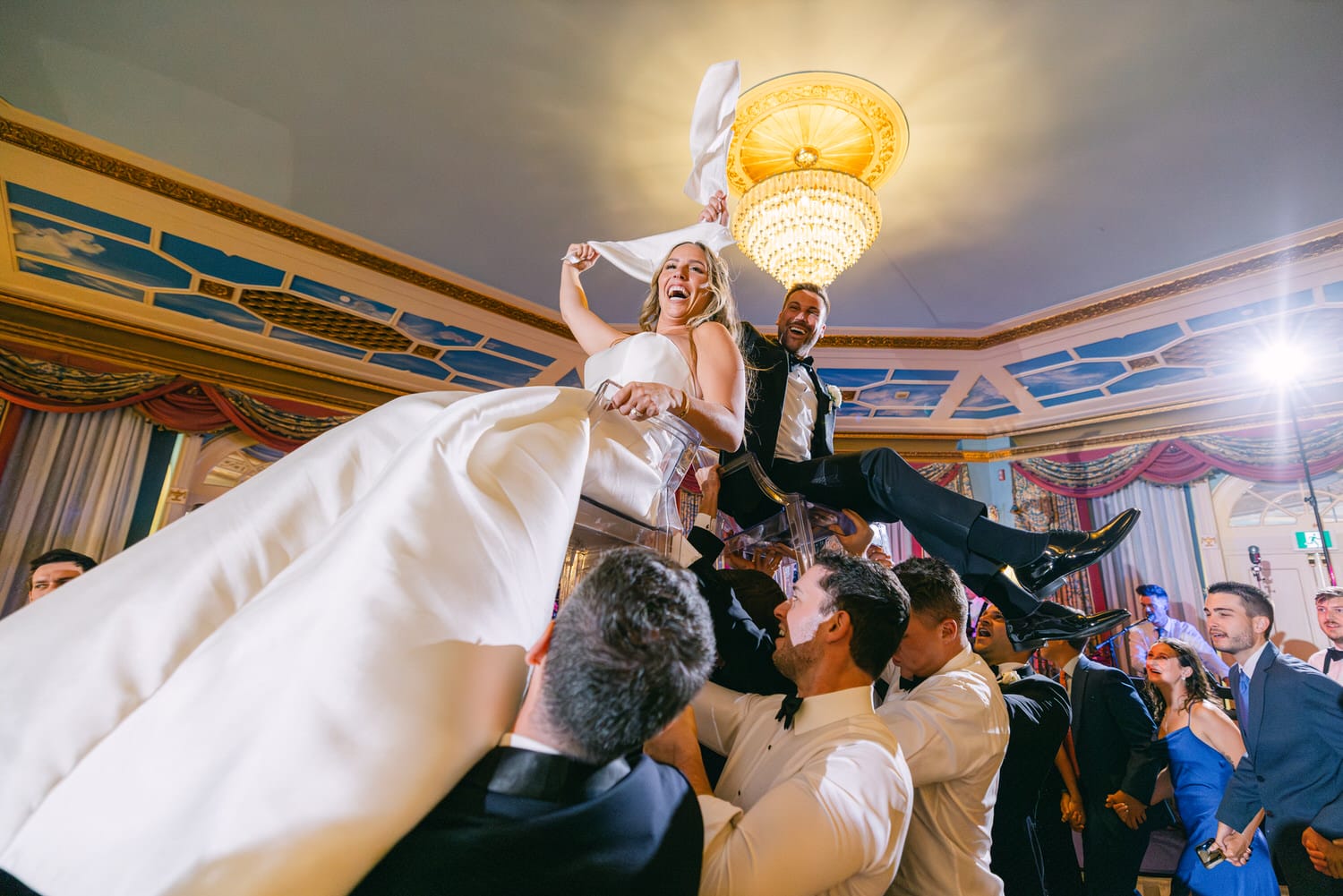 A joyful couple celebrates their wedding by being lifted by friends, surrounded by elegantly dressed guests and a luxurious venue.