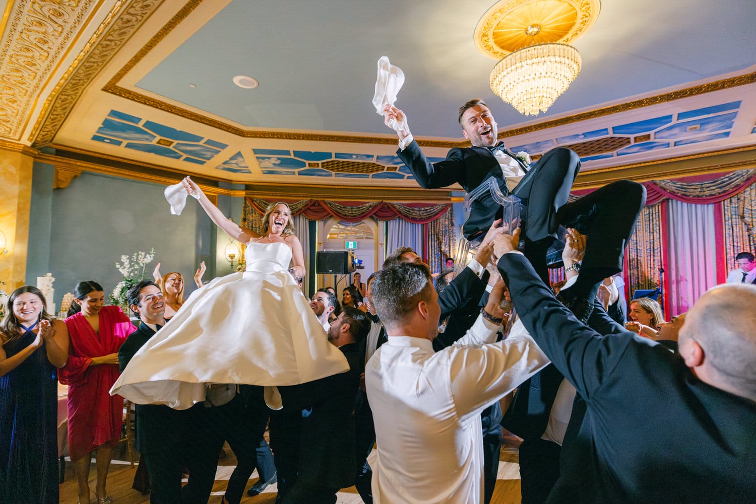 A joyful couple is lifted by guests during a wedding celebration, surrounded by friends and family in a beautifully decorated venue.