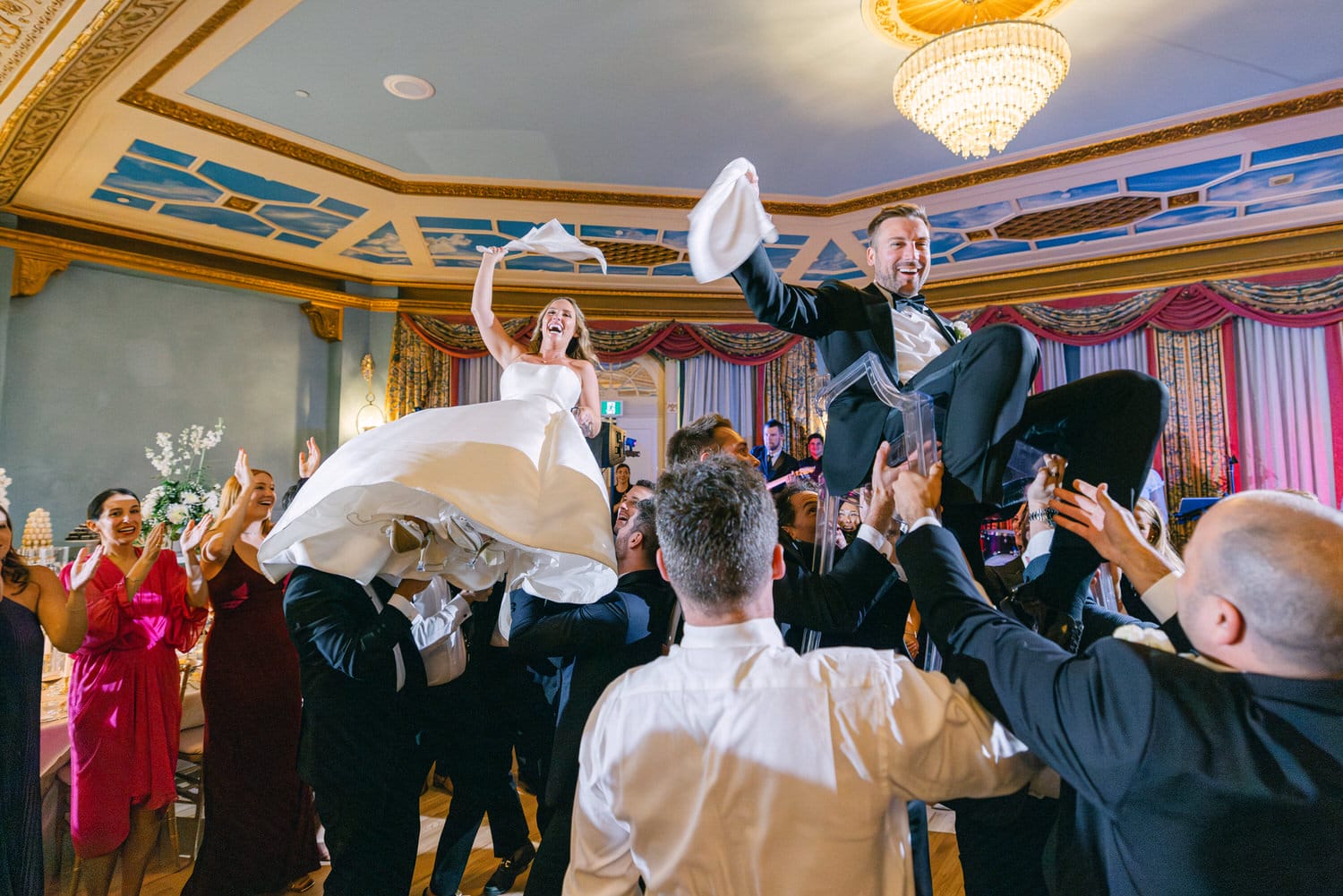 A joyful bride and groom are lifted by their guests during a lively dance at their wedding reception, surrounded by smiling friends and family.