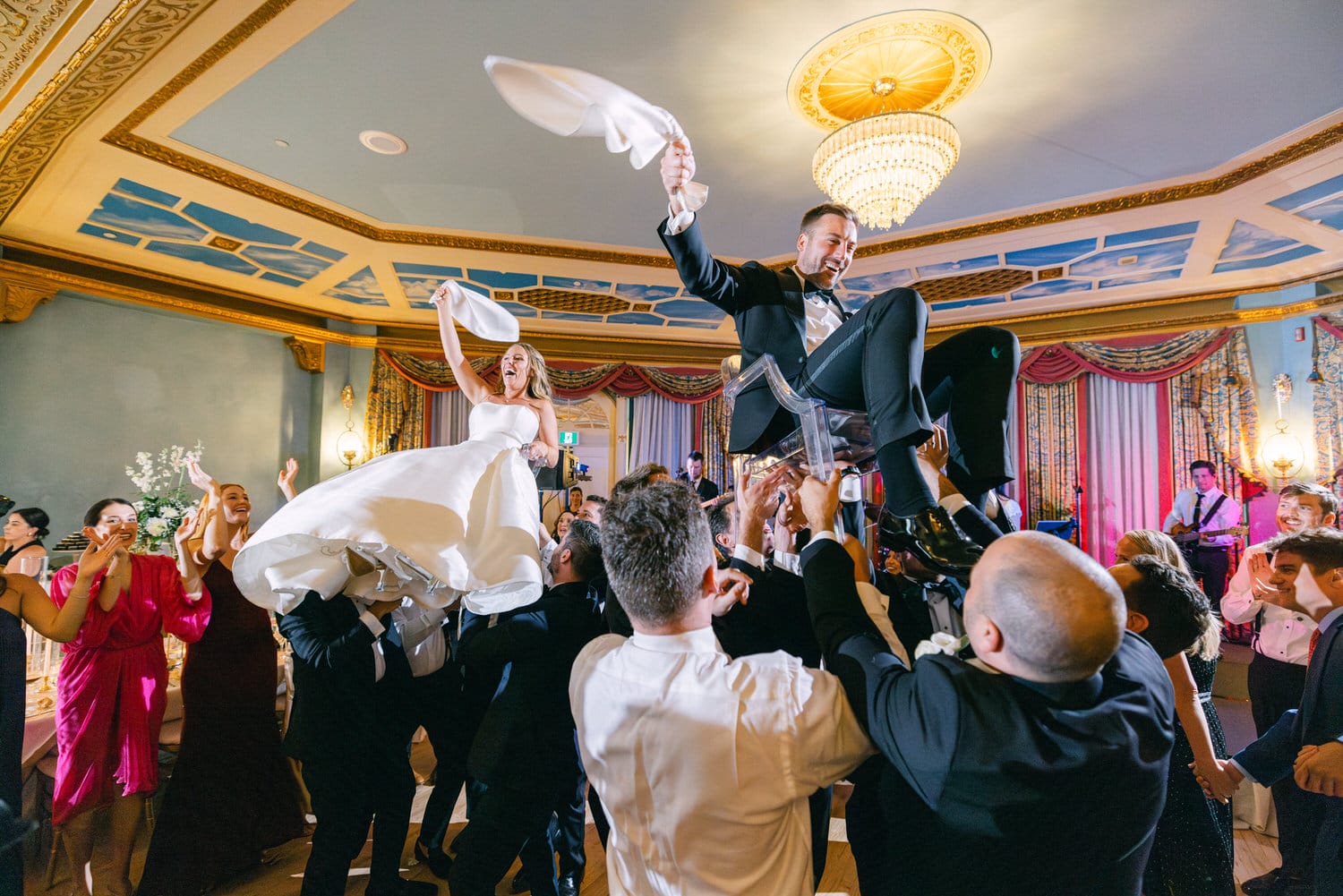 A lively scene of a wedding celebration where the bride and groom are joyfully lifted by guests, surrounded by vibrant energy and laughter.